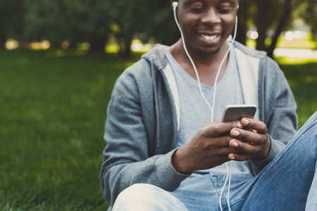 Smiling african-american student listening to music in earphones during break, sitting on the grass having rest in university campus outdoors. Education concept, copy spaceの写真素材