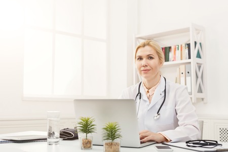 Portrait of happy female doctor sitting at the desktop. Physician is working in bright office, medicine concept, copy spaceの写真素材