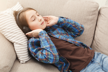 Cute girl listening to music in headphones, lying on beige couch, relaxing at home, copy spaceの写真素材