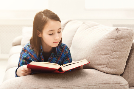 Cute little girl reading book. Dreamy casual child studying at home, lying on couch, copy spaceの写真素材