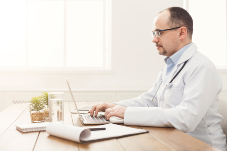 Serious doctor in glasses sitting at the desktop, typing on laptop. Physician is working in bright office, medicine concept, copy spaceの写真素材