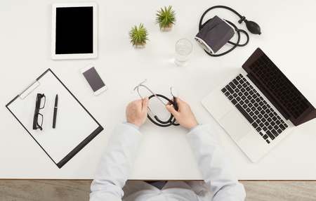 Doctor sitting at office desk with medical equipment all around and holding stethoscope, top view, copy spaceの写真素材