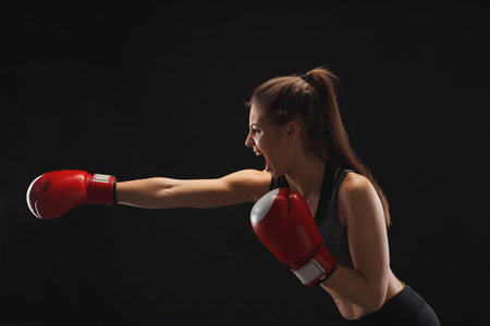 Side view of emotional young woman with boxing gloves and standing in position, ready to fight, copy space. Studio shot on black background, low key. Kickboxing and fight sport conceptの写真素材