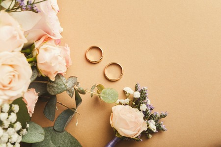 Wedding bride bouquet, floral corsage and two gold rings on beige background with copy space, top view. Preparation, wedding, advertisement, romance conceptの写真素材