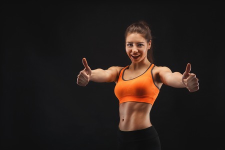 Athletic woman posing in studio and showing thumb up. Muscular body and strong muscles. Shot on black background, low key. Bodybuilding conceptの写真素材
