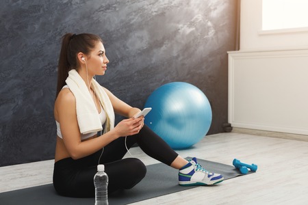 Happy fitness woman having rest and listening to music. Young girl sitting on mat in headphones. Bodybuilding, healthy lifestyle concept, copy spaceの写真素材