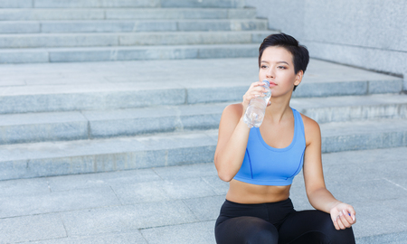 Young woman runner is having break, drinking water while jogging in city, sitting on staircase and looking away, copy spaceの写真素材