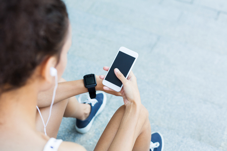 Image of an african-american woman using smartphone with blank screen outdoors, while listening to music, copy space, over shoulder shotの写真素材