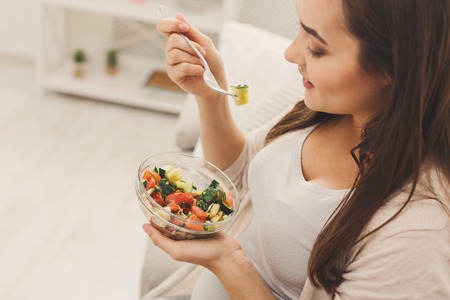 Young pregnant woman eating green salad. Attractive expectant lady sitting on sofa and having fresh snack. Healthy nutrition and pregnancy concept, copy spaceの写真素材