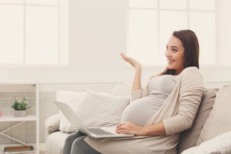 Excited pregnant woman using laptop, sitting on sofa at home. Young expectant lady surfing net, remote working, buying goods for newborn. Online shopping, pregnancy concept, copy spaceの写真素材