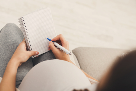Unrecognizable pregnant woman lying on sofa with blank notebook and pen and writing names for her baby, making shopping list, back view, copy spaceの写真素材