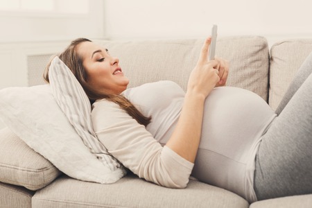 Happy pregnant woman using smartphone lying on sofa. Young expectant lady surfing the net, reading about baby care, buying goods for newborn. Online shopping, pregnancy conceptの写真素材
