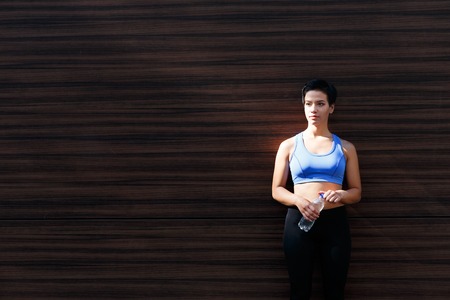 Young woman runner is having break, drinking water while jogging in city, leaning at dark wooden wall, copy spaceの写真素材