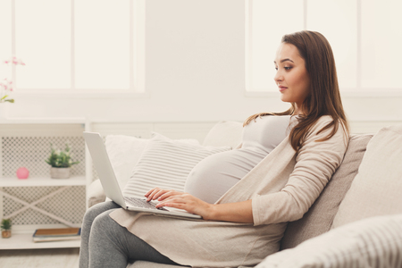 Pregnant woman using laptop, sitting on sofa at home. Young expectant lady surfing net, remote working, buying goods for newborn. Online shopping, pregnancy concept, copy spaceの写真素材