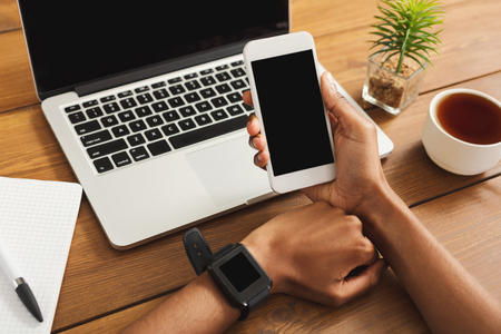 Black female hands using digital gadgets at office desktop. African-american woman synchronizing her smart watch, smartphone and laptop. Modern technology, blank screens for copy spaceの写真素材