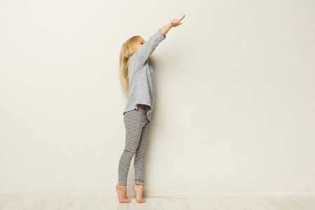 Full length portrait of a happy little girl standing and pointing aside with finger on white studio background, side view, copy spaceの写真素材