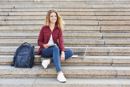 Young smiling female student sitting on the stairs using laptop outdoors during break, having a rest in the university campus. Technology, education and remote working concept, copy spaceの写真素材