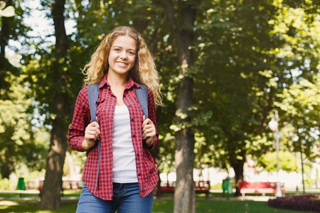 Happy young female student standing in the park with backpack behind her shoulders. Technology,...