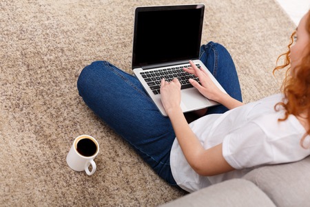 Unrecognizable young woman typing on laptop keyboard while sitting on the floor with coffee. Back shot, blank screen of computer for copy spaceの写真素材
