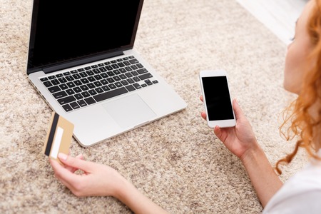 Unrecognizable young woman shopping online on laptop with credit card and phone while lying on the...