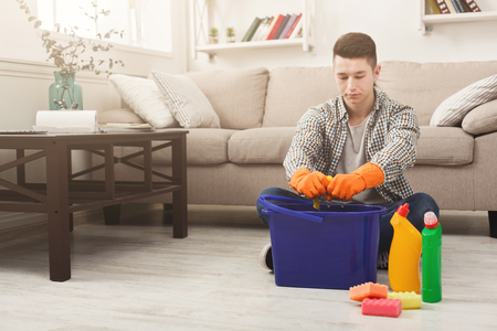 Tired man winkling out towel and cleaning flat with detergents in living-room. Housekeeping and home cleaning conceptの写真素材