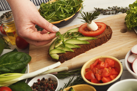 Female hands making avocado bruschettas with cheese, arugula and tomato. Healthy vegetarian sandwiches at kitchen table with various vegetables bowls and greens. Cooking food background, closeupの写真素材