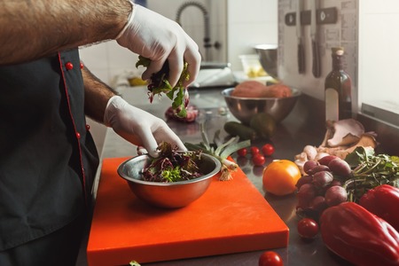 Unrecognizable male chef in gloves preparing vegetable salad, seasoning lettuce leaves with olive oil in professional hotel or restaurant kitchen interiorの写真素材