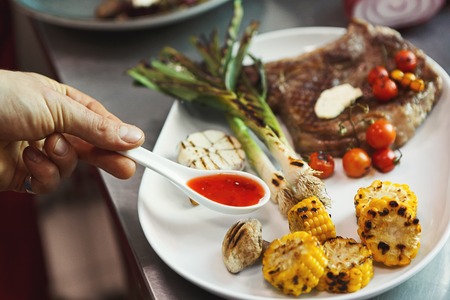 Male chef cooking and serving juicy medium beef steak with sauce and grilled vegetables. Healthy exclusive food prepared at professional kithenの写真素材