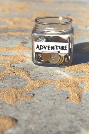 Glass jar with coins for adventure on gray floor with sand background, copy space. Money box, distribution of cash savings concept.の写真素材