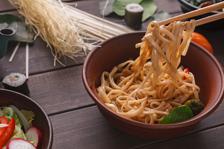 Chopsticks, taking rice noodles with soy sauce in bowl on wooden background, close-up.の写真素材