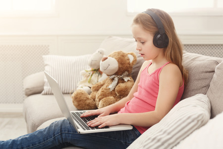 Little girl in earphones with laptop at home. Cute child doing homework on computer. Modern online...