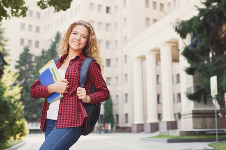 Beautiful smiling female student holding notebooks in hands while standing in front of the university campus. Education concept, copy spaceの写真素材