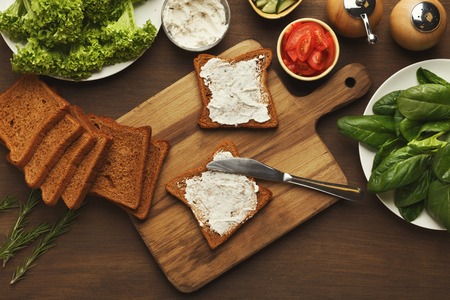 Healthy food preparation. Top view on wooden cutting board, wholegrain rye bread with soft ricotta cheese topping, cut organic tomato, olives and greens, copy spaceの写真素材