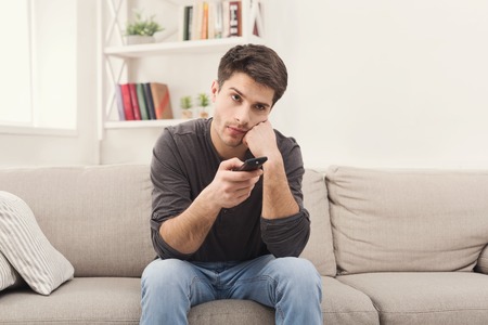Young man watching television, using remote control to switch channels. Guy bored with what he sees on TV screen. Sitting on couch in living room at home, copy spaceの写真素材