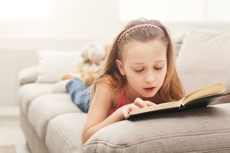 Cute little girl reading book while lying on sofa at home. Education and leisure concept, copy space on windowの写真素材