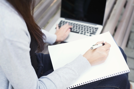 Unrecognizable student girl sitting on bench in park, working with laptop and taking notes, preparing for exams outdoors, having rest in university campus, copy spaceの写真素材