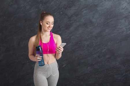 Happy fitness girl in earphones listening music on her smartphone and drinking water after training. Modern technologies, healthy lifestyle conceptの写真素材