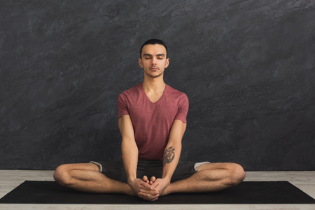 Young man practicing yoga, meditation exercises. Guy does lotus pose for relaxation, sitting with closed eyes on mat at gym. Healthy lifestyle, copy spaceの写真素材