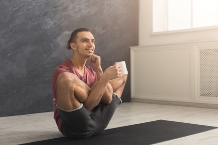 Young smiling man in flexible yoga pose drinking tea, sitting on mat at gym and practicing twisting exercise, copy spaceの写真素材