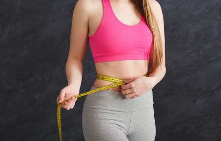 Unrecognizable fit woman measuring her waist on grey background. Happy sporty girl with measure tape. Weightloss, healthy lifestyle, bodycare conceptの写真素材