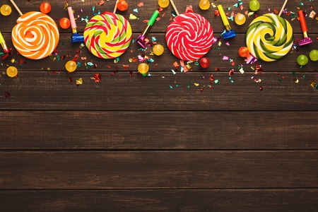 Border of colorful spiral lolipops, confetti and small bon-bons on rustic wood. Birthday party decoration background, top view, copy space. Mockup for greeting cardの写真素材