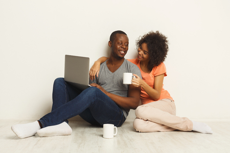 Happy black couple shopping online, making order with laptop while sitting at home floor in empty living room, copy space, isolatedの写真素材
