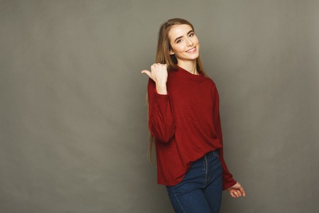 Young confident happy woman point away. Attractive girl in red pullover gesturing with her finger aside, studio background, copy spaceの写真素材
