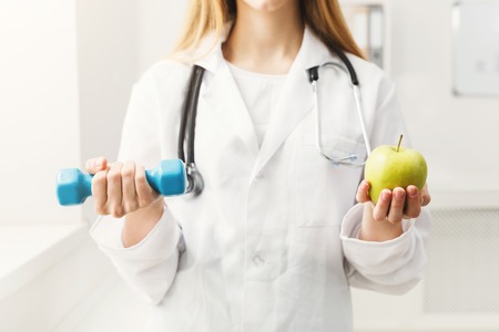 Young unrecognizable dietitian with apple and dumbbell at clinic. Woman nutritionist holding fruit and sport equipment. Weight loss, healthy eating and slimming conceptの写真素材