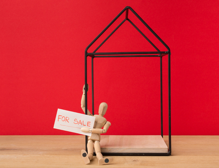 House for sale on red background. Wooden mannequin holding advertisement board. Real estate and buying from owner concept, copy spaceの写真素材