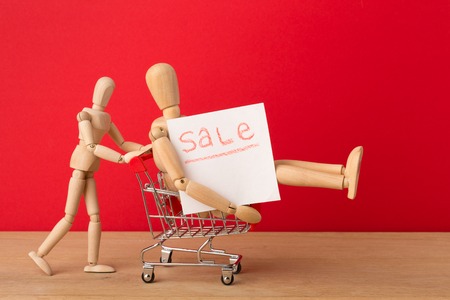 Wooden mannequins with miniature shopping cart running for sales on red background. Mockup for shopping, discount, black friday, copy space, side view.の写真素材