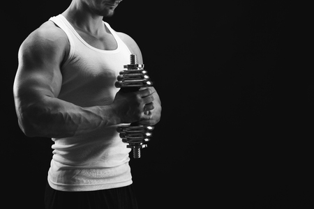 Athletic young man with dumbbell, black and white image. Muscular body and shoulder muscles. Studio shot, low key. Bodybuilding concept, copy space, cropの写真素材