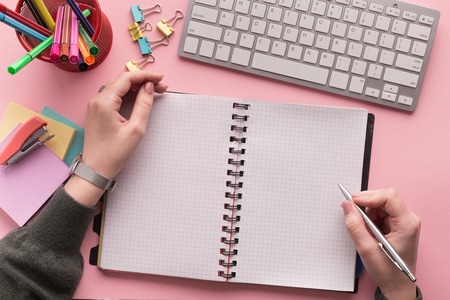 Womans hands writing in spiral notepad as mockup for your design on pink background with keyboard and stationery, top view. Girlish workplace, lifestyle concept with copy spaceの写真素材