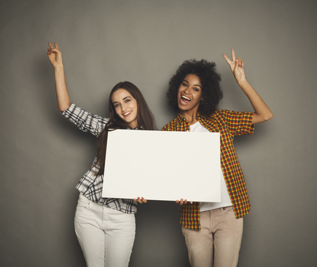Multiethnic girls holding blank white banner with copy space on gray studio backgroundの写真素材