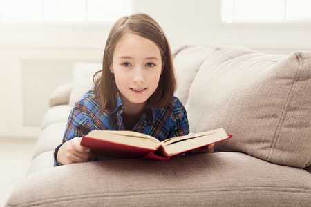 Pensive girl reading book. Young cute woman studying at home, lying on couch, copy spaceの写真素材